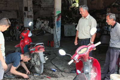 Drei Männer stehen um einen am boden knieenden Mechaniker der ein Mofa repariert