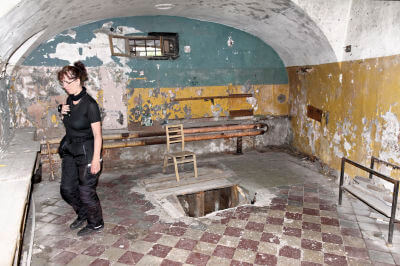 Frau steht im Exekutionsraum dahinter ein Loch im Boden mit einem Holzstuhl dahinter