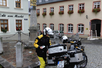 Motorradfahrerin steht bei Regen am Marktplatz vor der Postmeilensäule in Wolkenstein.