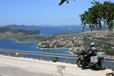 Motorradfahrer vor Leitplanke dahinter blaues Meer mit Bergen