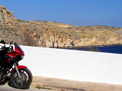 Motorrad parkt vor einer weißen Mauer und dahinter Felsen und das Meer bei den Dingli Cliffs