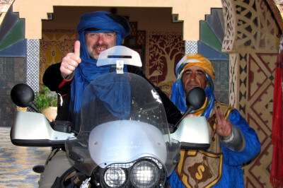 Ein Motorradfahrer auf seinem Motorrad sitzend und mit Turban ausgestattet, daneben ein Beduine in tradtioneller Kleidung im Hotel Kasbah Asmaa