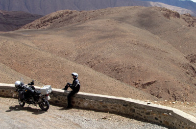 Frau sitzt neben Motorrad auf einer kleinen Steinmauer auf der Passhöhe des Tizi-n-Tirherhouzine