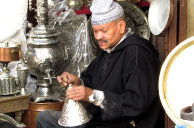 Ein im schwarzen Gewand gekleideter Kupferschmied bearbeitet eines seiner Produkte