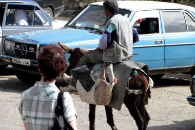 Mann auf bepacktem Esel vor einem blauen Mercedes Taxi