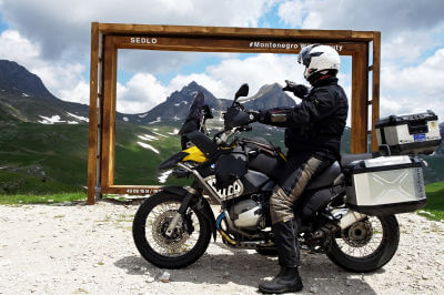 Motorrad mit Fahrer steht vor Holzrahmen und Zeigt mit Hand auf die Doppelgipfel des Sedlena Greda