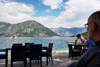 Mann sitzt in einem Kaffee und blickt über Stühle und das Meer auf Berge