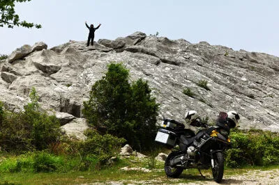 Frau steht auf Felsen und winkt und unten parkt ein Motorrad