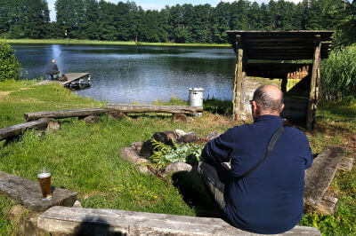 Mann sitzt auf kleinem Holzbänkchen und Blick an einer Holzhütte vorbei auf den an die Unterkunft angrenzenden See