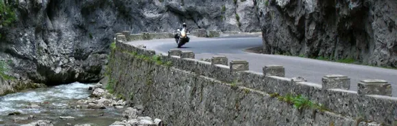 Motorrad fährt auf schmaler Straße mit Randbegrenzung aus Stein in einer Schlucht an einem Fluss entlang