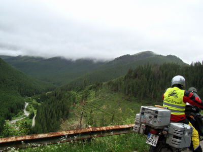 Motorradfahrer mit gelber Warnweste steht an Leitplanke und blickt hinunter ins bewaldete Tal