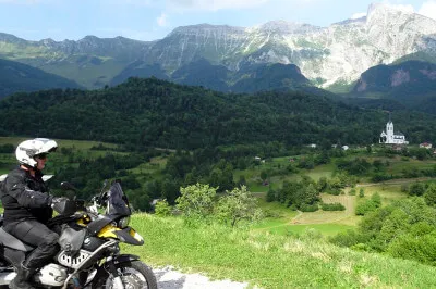 Eine kurvige Straße führt von Kobarid hinauf nach Drežnica, dahinter ragt unübersehbar der Krn auf. Motorradfahrer vor Bergkette mit Dorf im Hintergrund