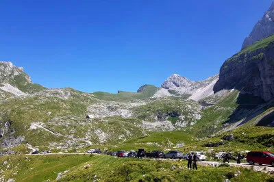 Ein kurzer Stich führt noch hinauf bis zur Mangarthütte, Koca na mangartskem sedlu, auf 1906 Metern. Wer ein Bett oder Verpflegung sucht, wird hier fündig. Rechts der Hütte ragt der Gipfel Mangart in den Himmel. Gipfelbereich mit Blick auf parkende Autos und einer faszinierenden Berglandschaft auf der Mangartstraße