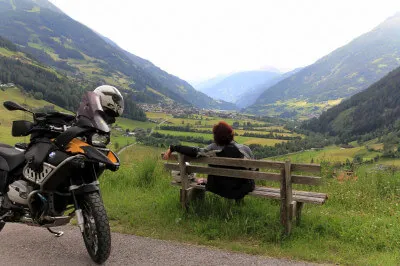 Wir sitzen am Rand einer kleinen Straße auf einer Bank und genießen den Blick hinunter auf's Matreier Becken, hinter uns das hoch aufragende Tauerngebirge. Frau mit Motorrad auf Holzbank mit Blick ins Tal