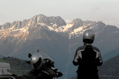 Motorradfahrerin steht auf der Göscheneralp und blickt auf schneebedeckte Berge