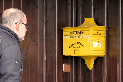 Mann steht vor am Haus hängenden gelben Briefkasten beim Misthaus