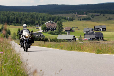 Motorrad fährt auf Straße und im Hintergrund ein verstreutes Anwesen in Jizerka